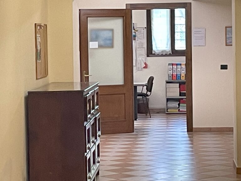 Corridoio con mobile e bacheca, porta aperta su stanza ordinata con scrivania e raccoglitori